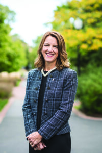 President Mary Surridge in blue plaid dress jacket and pearl necklace, standing at center of campus.