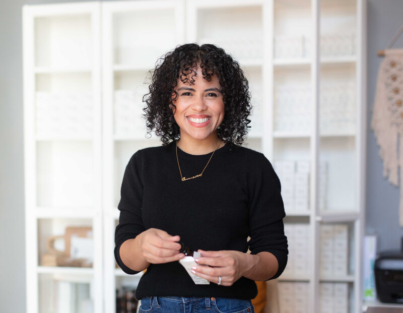 Woman stands in sunlit workspace holding a bottle of essential oil.