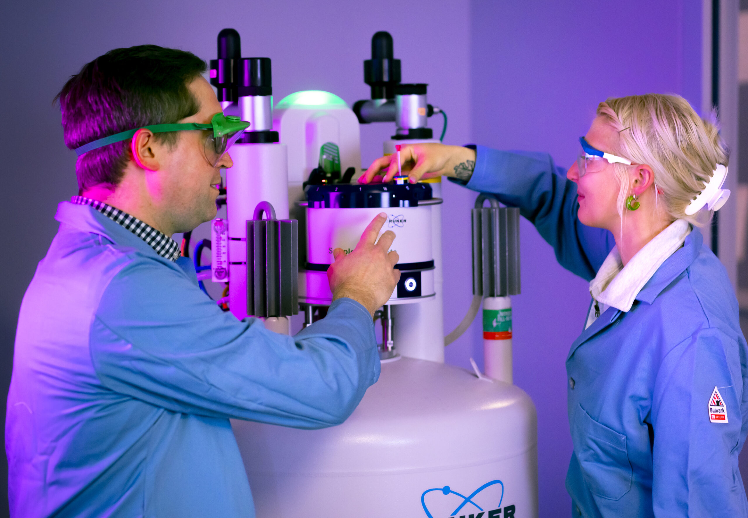 Two people in lab coats and safety glasses operate a Bruker scientific instrument in a laboratory setting, adjusting controls and discussing. The room is illuminated with a mix of blue and purple lighting.