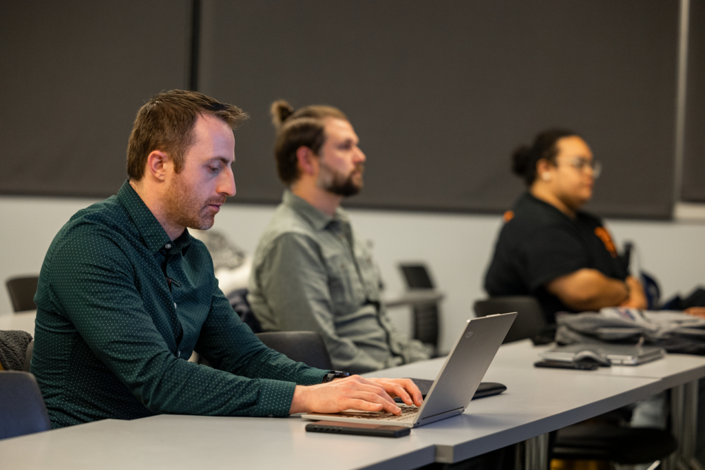 Three individuals sit in a classroom setting, focused on their own tasks. The nearest person types on a laptop, with the others listening attentively. The room is well-lit, and the atmosphere is studious.