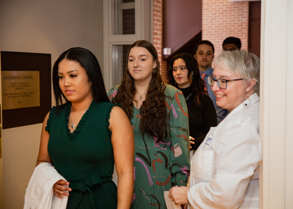 A group of nursing students walking in a hallway for their white coat ceremony, with a professor welcoming them into the chapel smiling.
