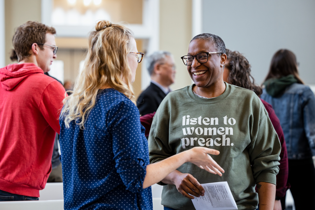 Two people are engaged in conversation in Isaacson Chapel. One is wearing glasses and a sweatshirt with the slogan 