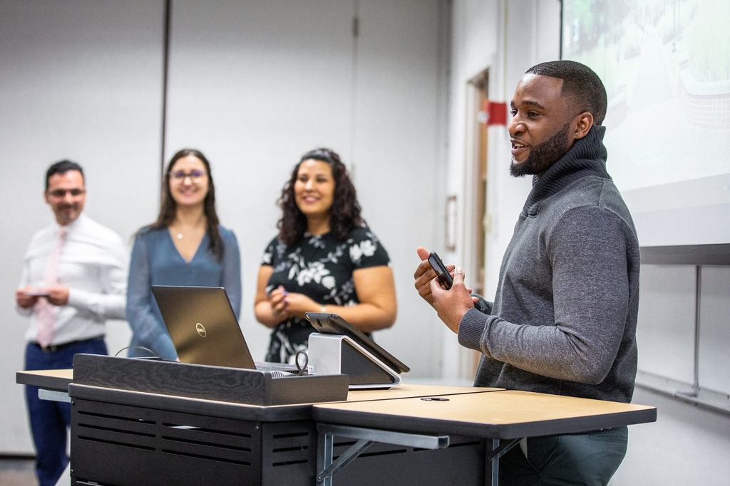A person stands at a podium with a laptop, speaking to an audience. Three individuals in the background are smiling while facing the speaker. The setting is a classroom.