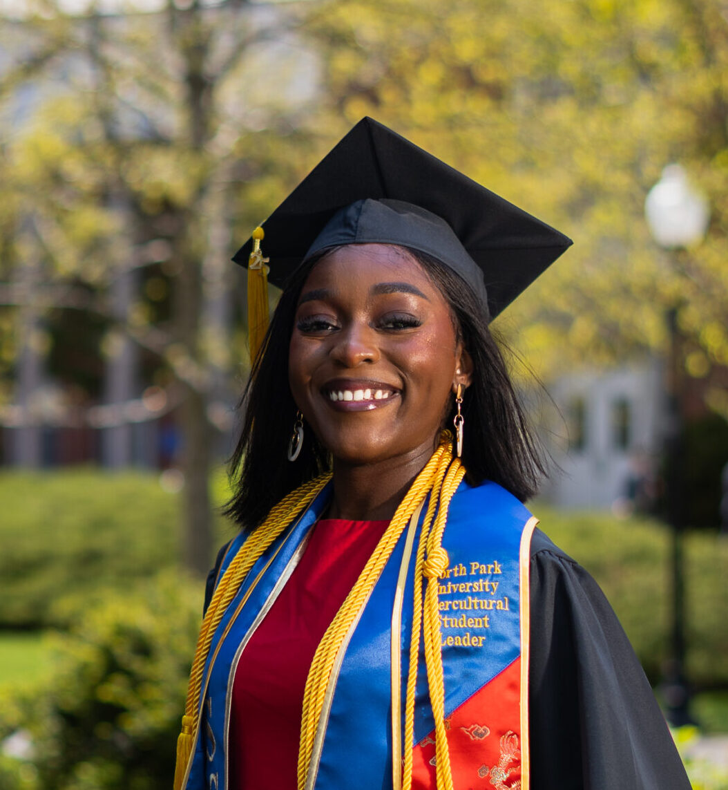 Image of Fidelie Futi wearing a graduation cap and gown, with honor cords and a sash from North Park University, stands outdoors.