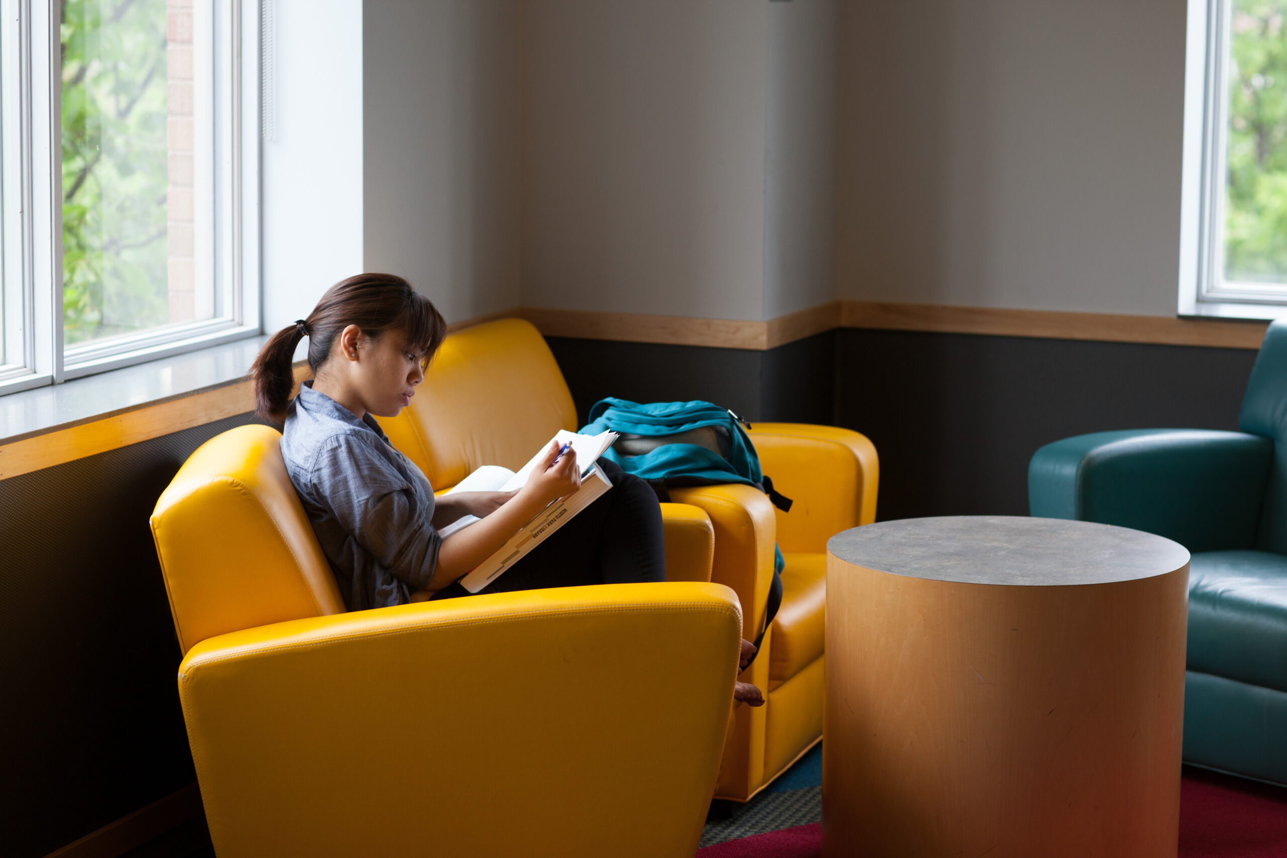 A person sits on a yellow chair beside a large window, engrossed in reading a book in Brandel Library. A turquoise backpack rests on the seat next to them. A round wooden table and another chair are nearby in the room.