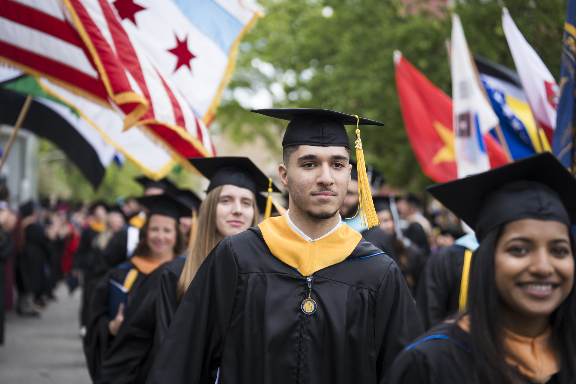 A group of graduates in black caps and gowns walk in a procession. One individual in focus wears a yellow tassel. Many flags, including the Chicago flag, are displayed in the background. Trees line the procession route.