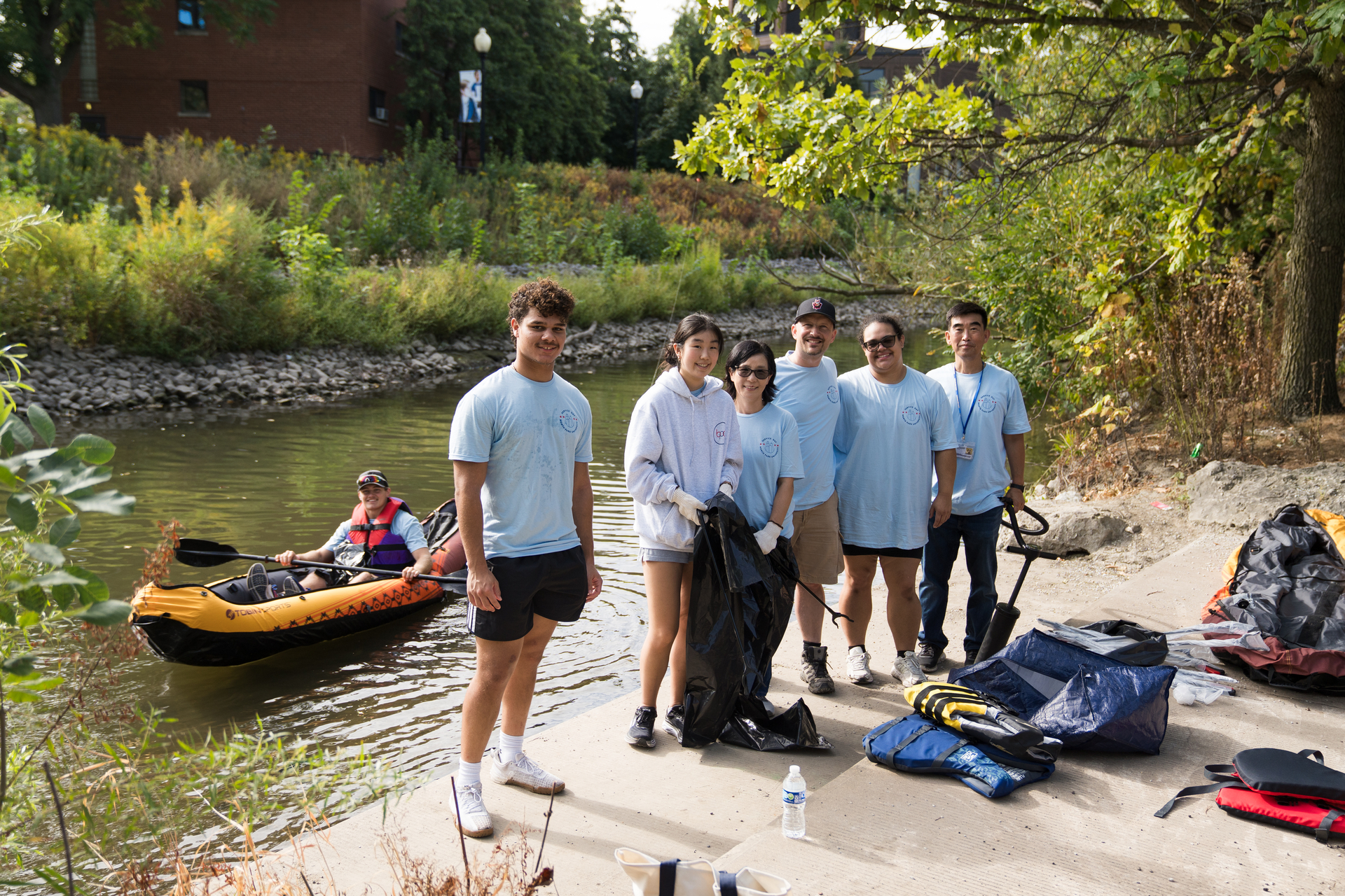 A group of people in light blue shirts are participating in a river cleanup for North Park University's annual Service Day, standing on a riverbank surrounded by trees and foliage. They are holding trash bags and tools. A person paddles in a small yellow inflatable boat on the water nearby. Various cleaning supplies and life vests are on the ground.
