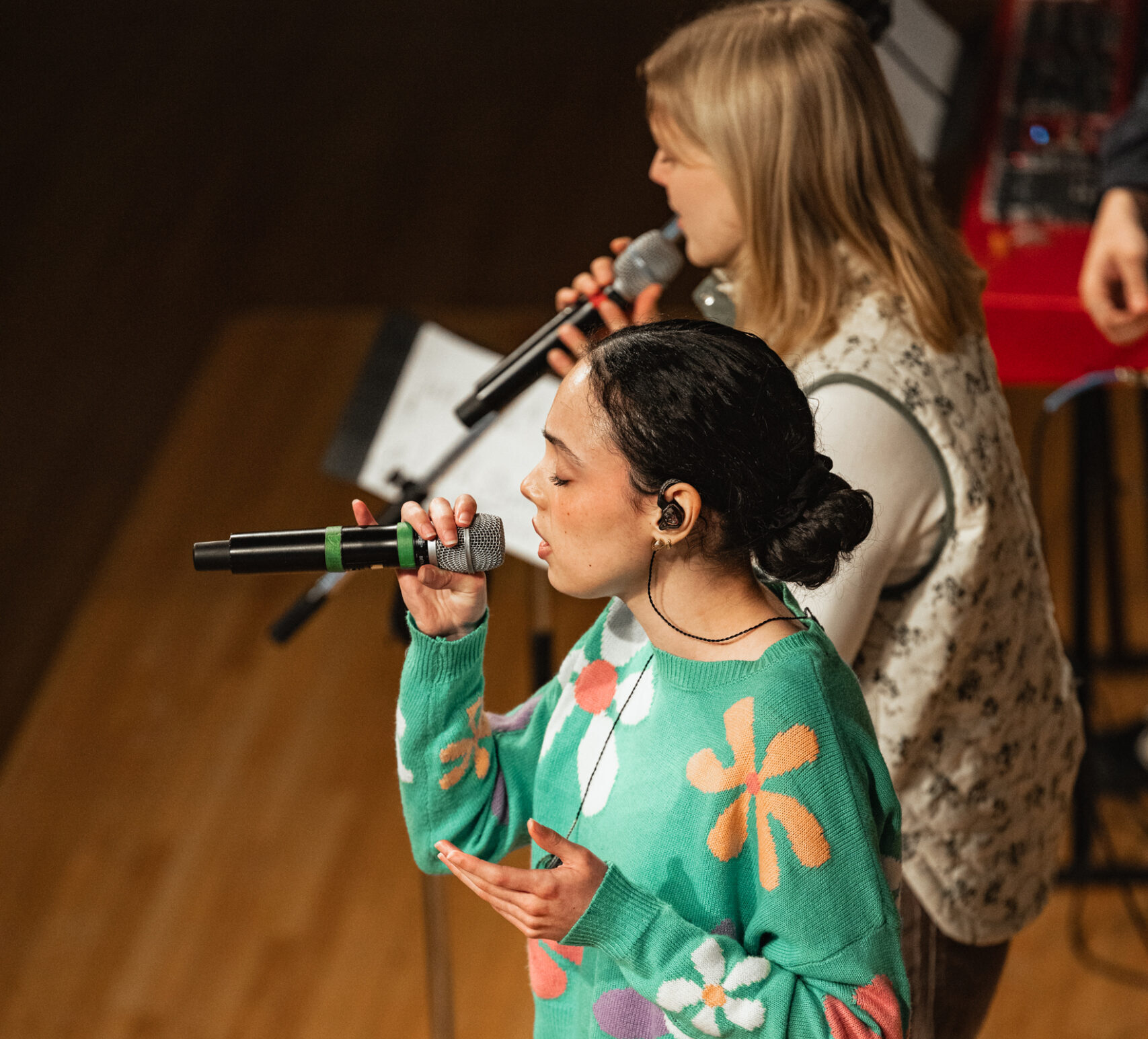 Two people singing on stage, each holding a microphone. The person in the foreground is wearing a green sweater with colorful flowers. A piano and sheet music are visible in the background.
