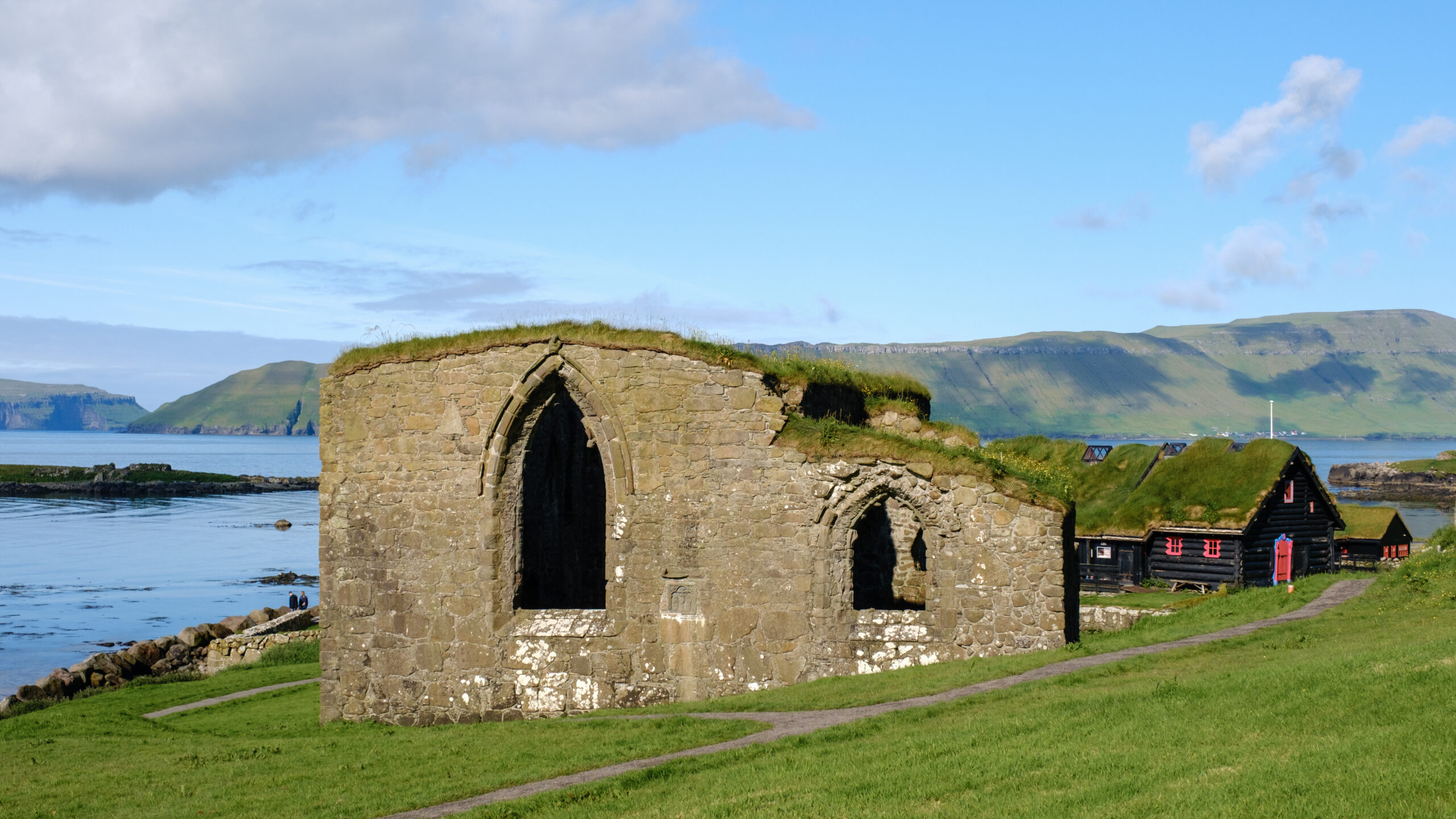 Ancient stone ruins with large arched windows stand in a grassy field near the coast on the Faroe Islands. Rolling hills and a blue sky are in the background.