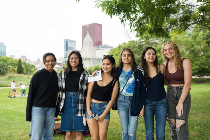 A group of young women stand together outside in a park in downtown Chicago, smiling at the camera.