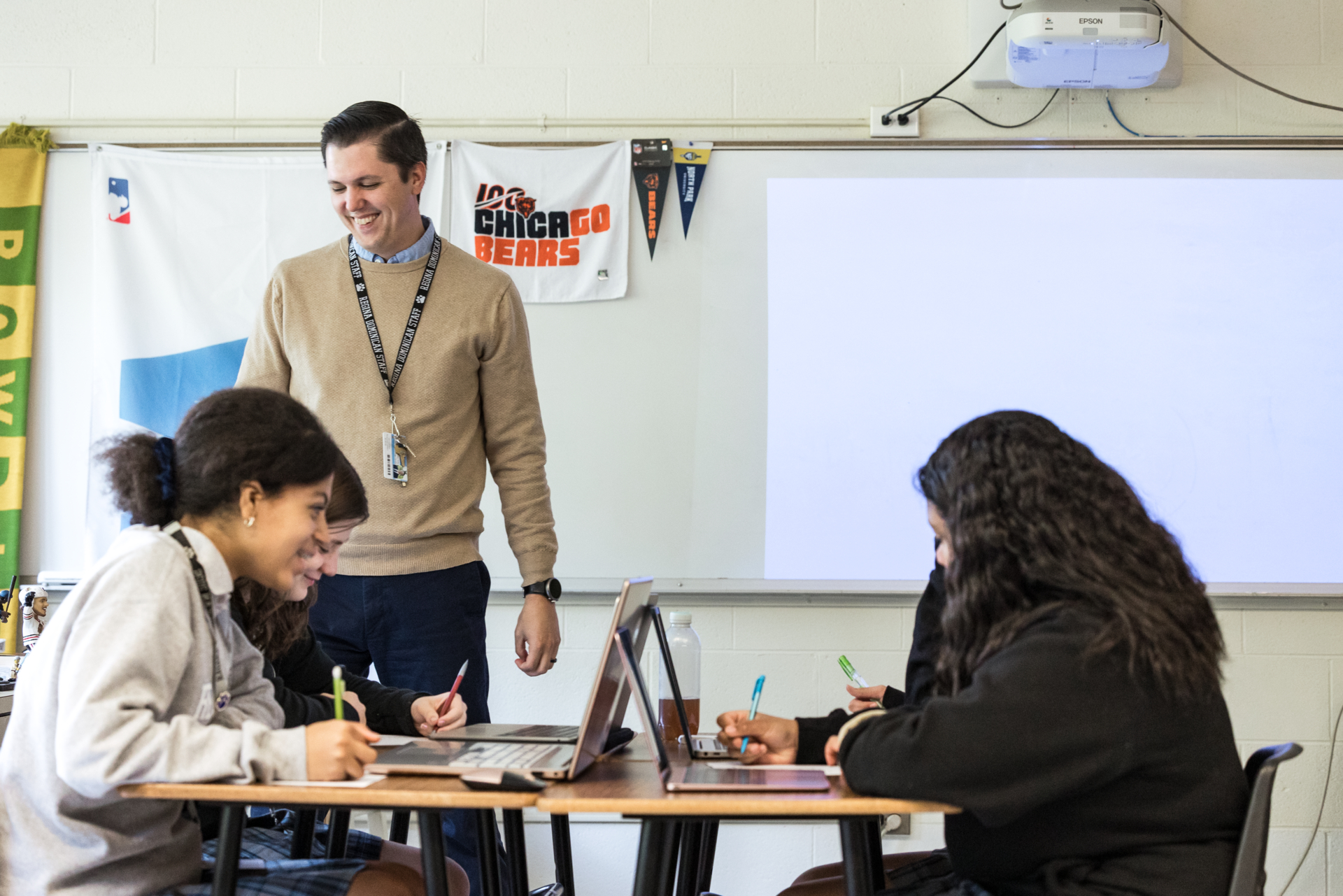A teacher stands at the front of a classroom, smiling, wearing a lanyard. Four students are seated at a table, working on laptops and writing notes. A 