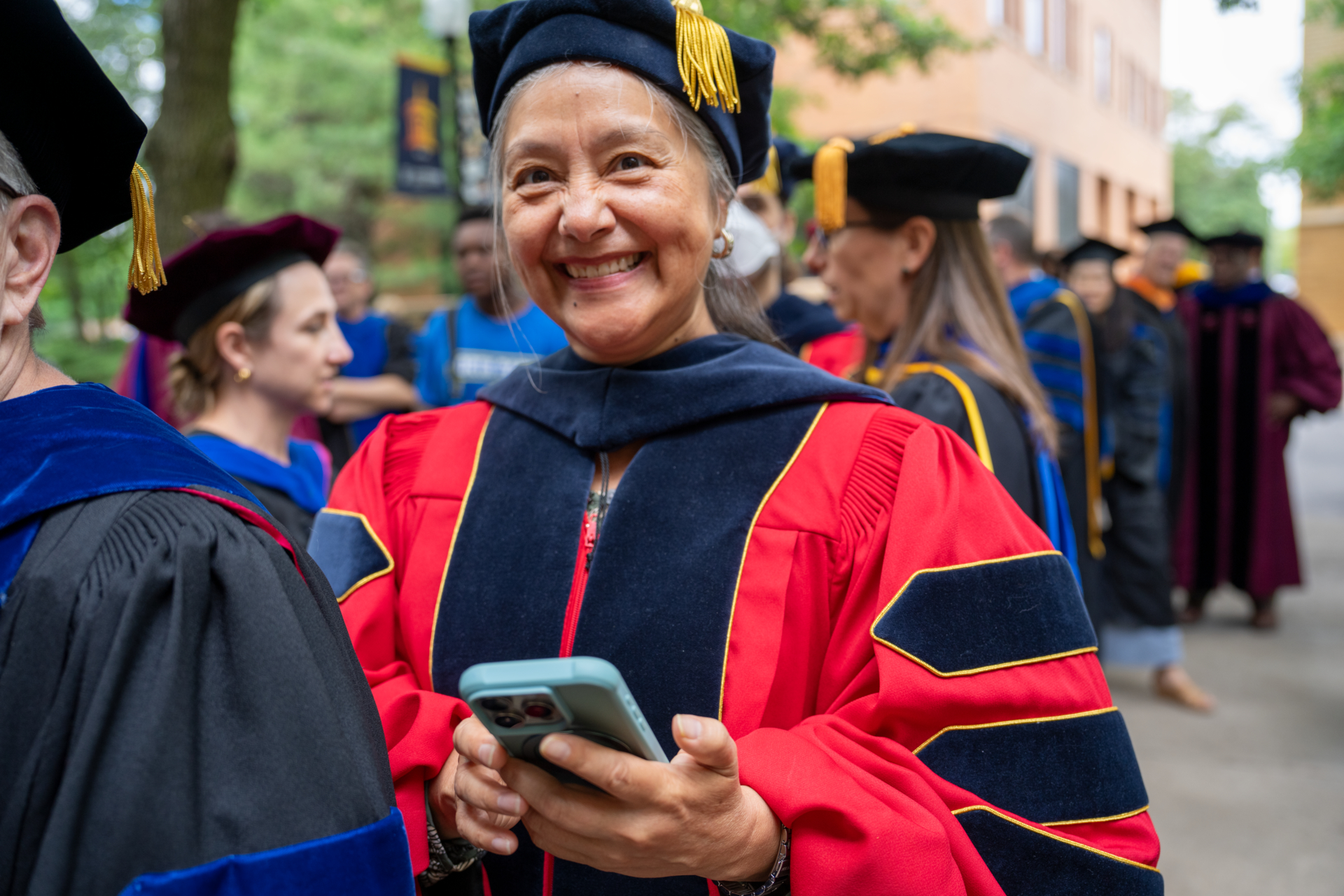 A professor in academic regalia is smiling and holding a phone during a graduation ceremony. Other individuals in similar attire are visible in the background. The setting is outdoors near a brick building.