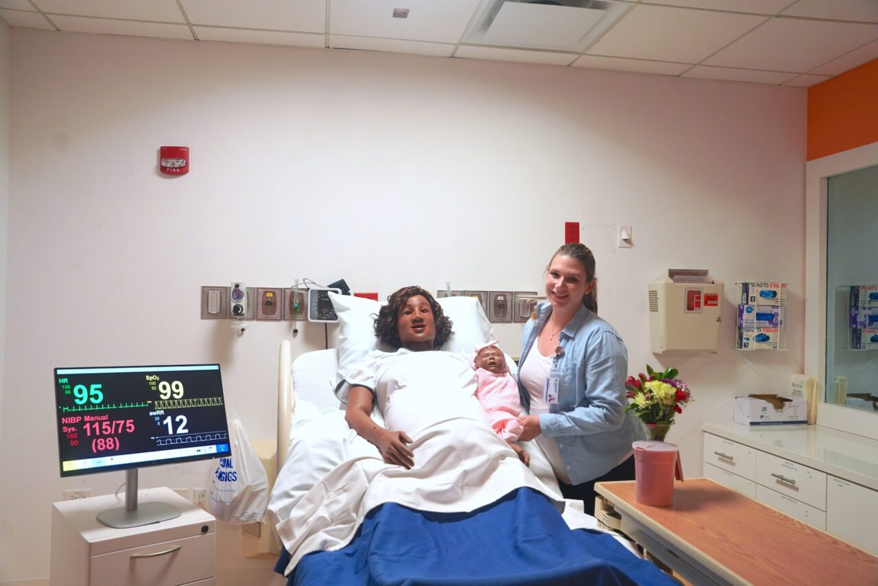 The simulation lab director stands in North Park's nursing lab next to a hospital bed holding a SimNewB infant mannequin. A Laerdal Medical MamaAnne birthing simulator is in the bed.
