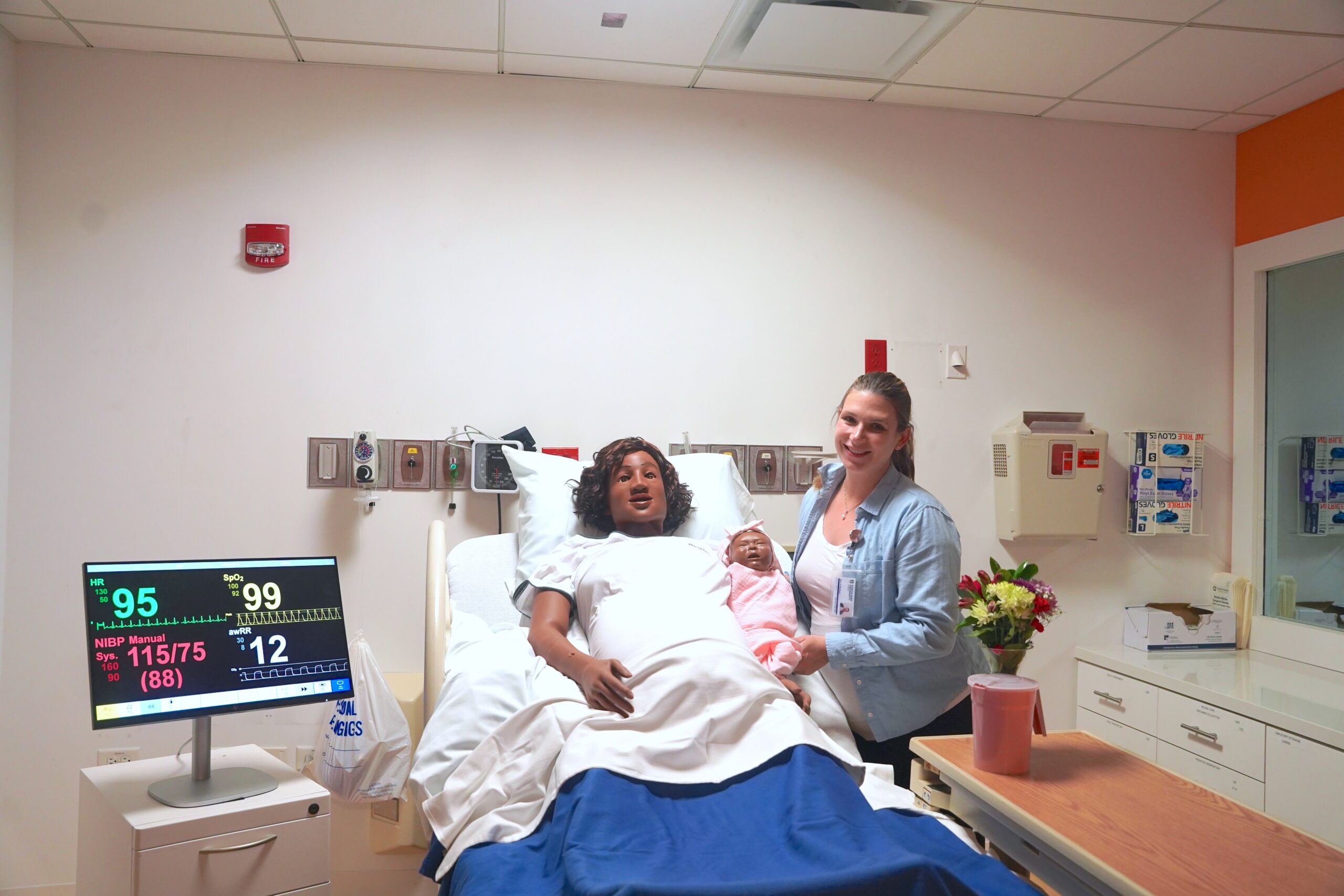 The simulation lab director stands in North Park's nursing lab next to a hospital bed holding a SimNewB infant mannequin. A Laerdal Medical MamaAnne birthing simulator is in the bed.