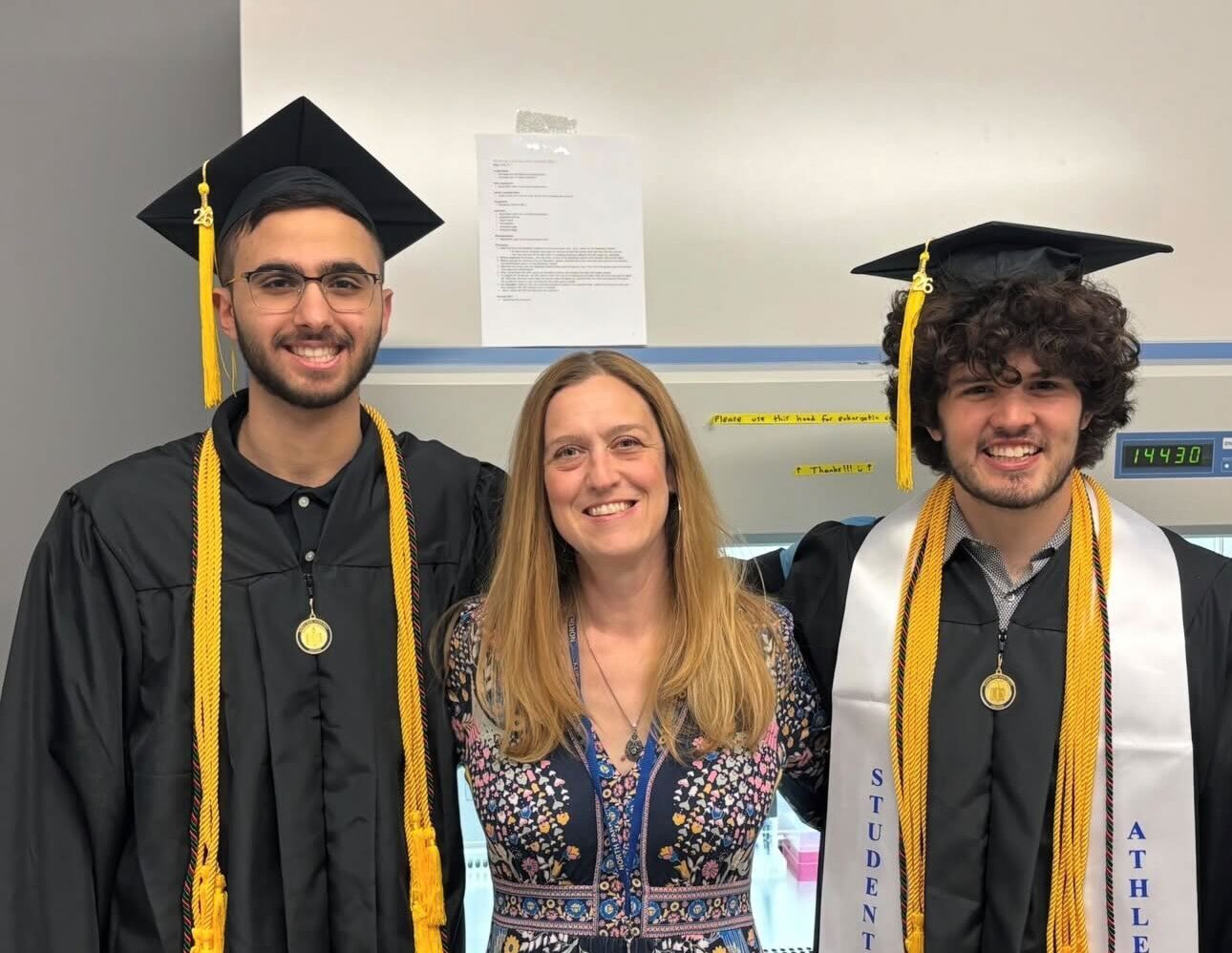 Two students in their graduation cap and gowns with one of their professors and advisors between them. All are smiling.