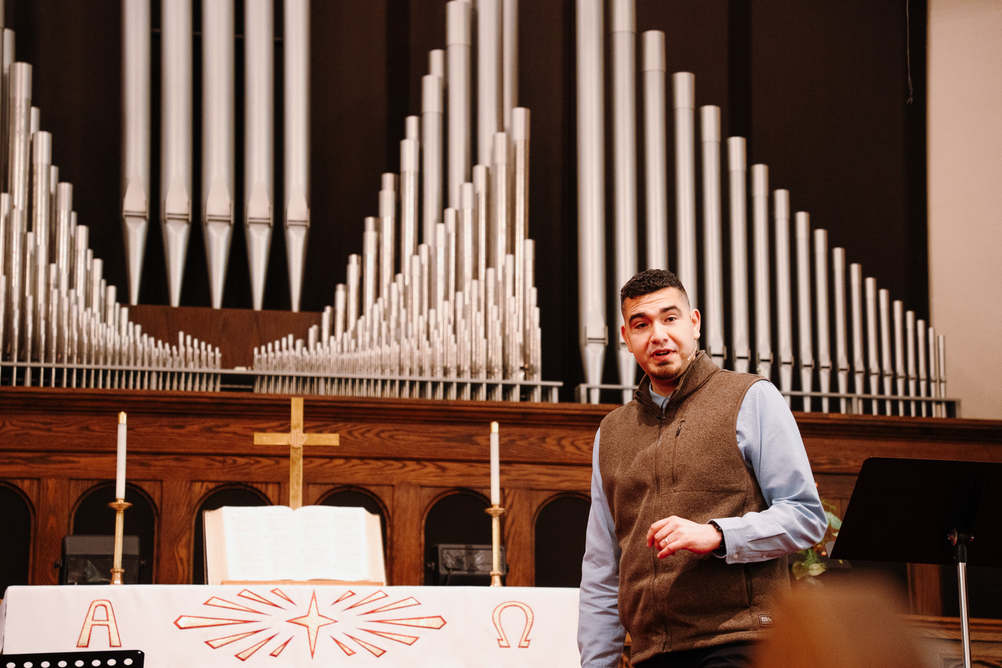 A man stands on an alter in a church, preaching. There is an organ behind him, as well as a cross and candles.
