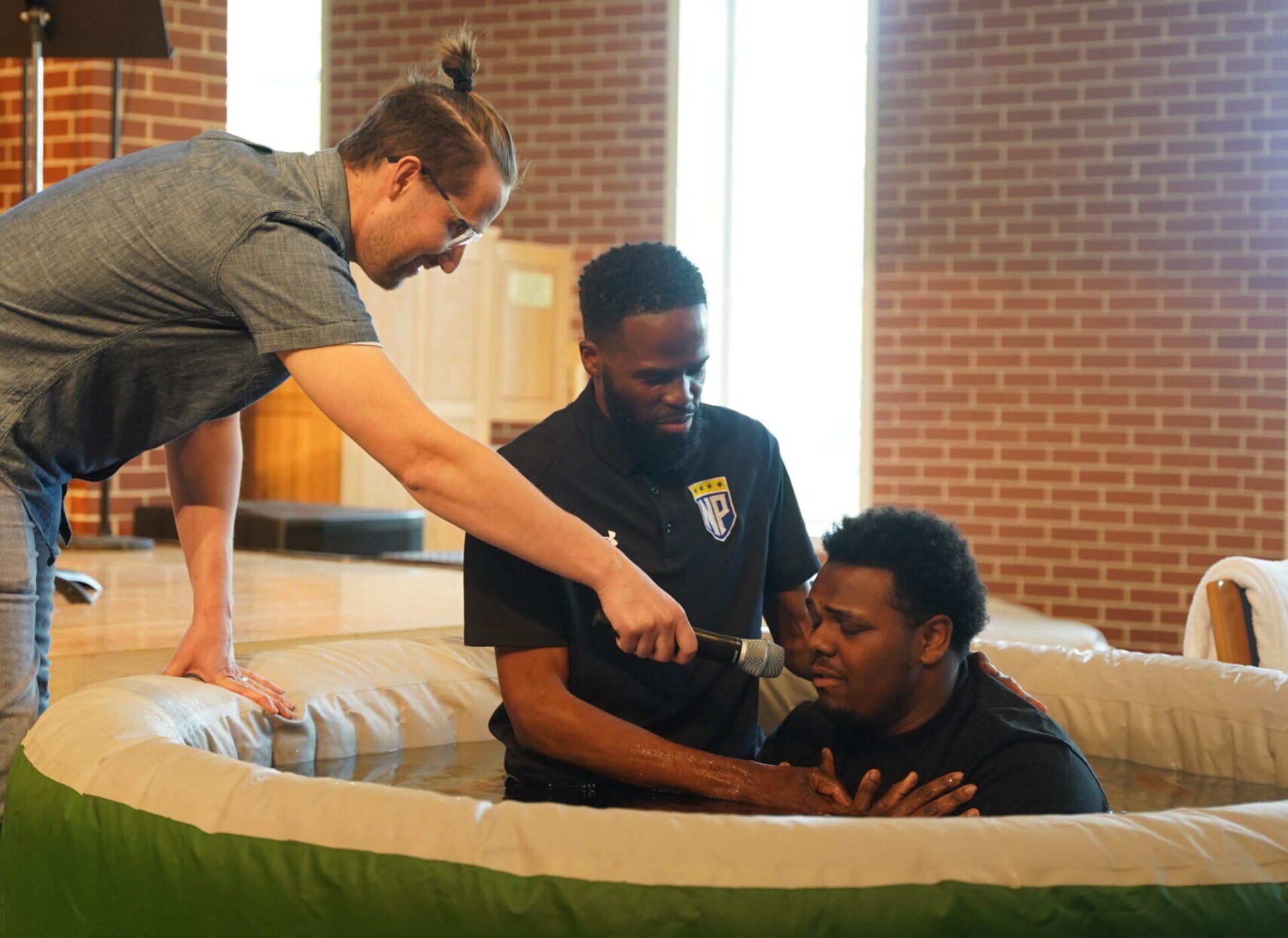 A pastor baptizes a student in an inflatable pool in the campus chapel. Another minister holds a microphone near the student's mouth.