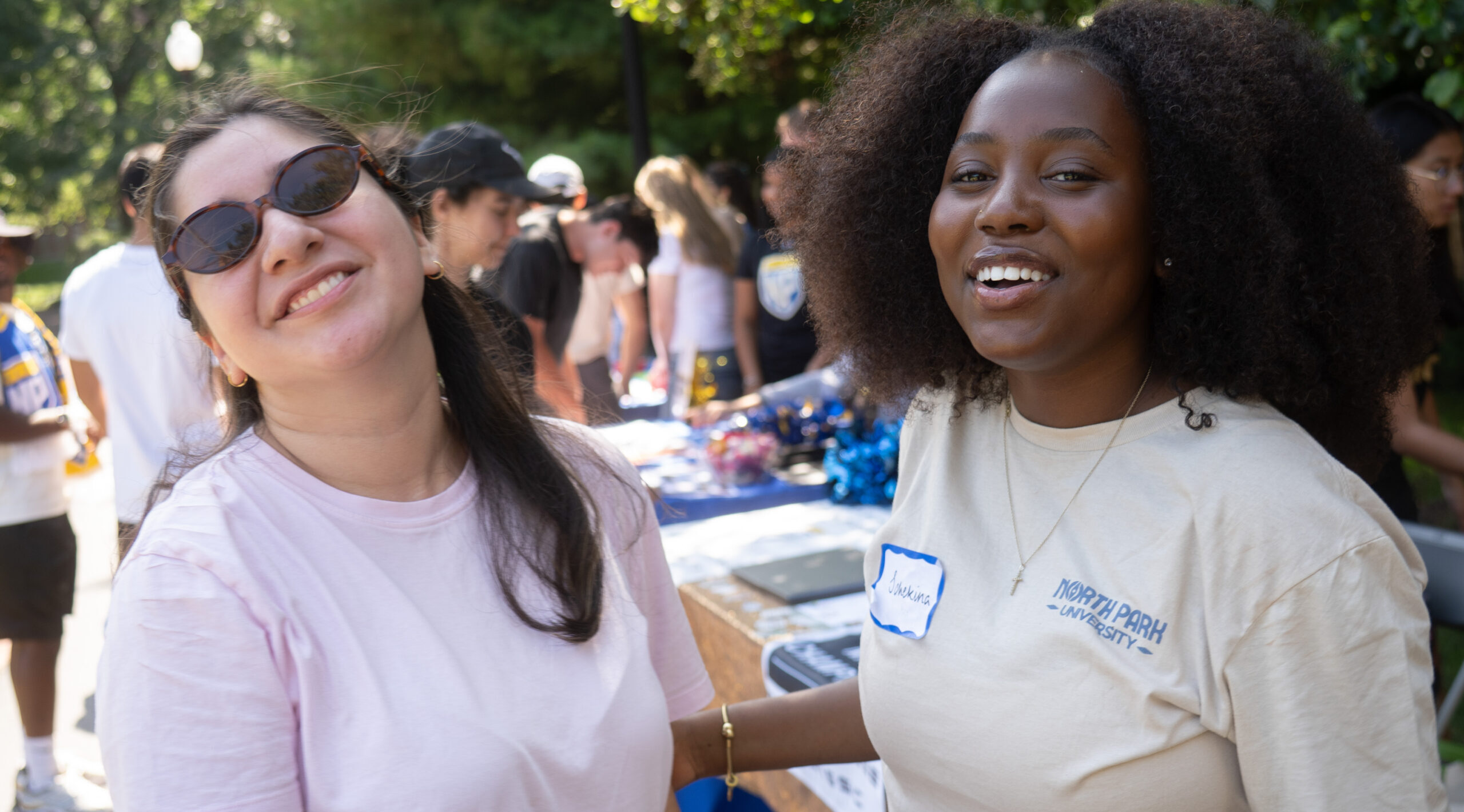 Two North Park students at Threshold New Student Orientation smile at the camera. One has sunglasses on, and they are outside on campus.