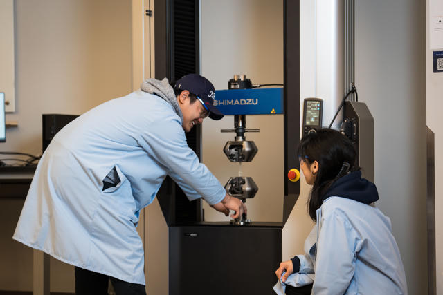 Two students in lab coats stand over a machine smiling as they work